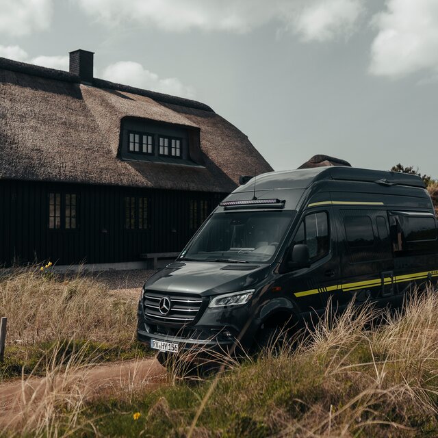 Schwarzer Camper Van mit grüner Beklebung steht seitlich in den Dünen vor einem braunen Haus, welches im dänischen Stil gebaut ist