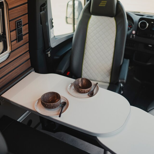 Gedeckter Tisch in einem modernen Camper Van, die Fahrersitze mit grau schwarzem Leder sind zum Tisch gedreht, links unten im Bild sieht man die Sitgruppe und links oben das Seitenfenster