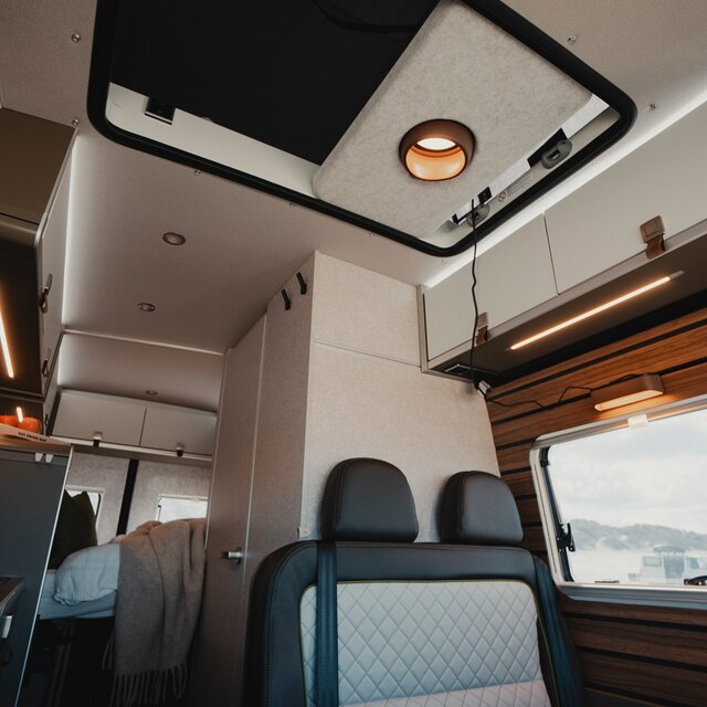 Wohnraum in einem modernen Camper Van mit Blick Richtung Decke, Rückenlehne der Sitzgruppe, Dachstauschränke und das Außenfenster sind zu sehen sowie der offene Durchgang an der Decke der zum Schlafdach führt, links im Bild sieht man die Küchenzeile mit Kochutensilien und im hinteren Bereich des Camper Vans sieht man das gemachte Heckbett
