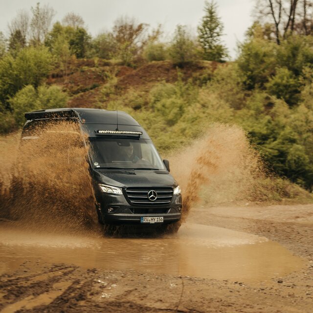 Camper Van fährt durch eine große Pfütze in einem Offroad Park, Pfütze spritzt, im Hintergund sind Bäume und Dreckhügel zu sehen