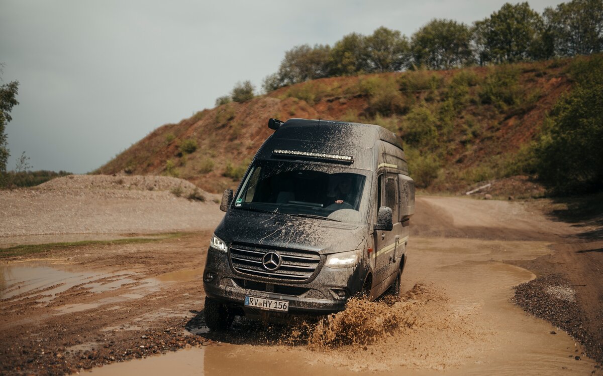 Camper Van fährt durch eine kleine Pfütze in einem Offroad Park, im Hintergund sind Bäume und Dreckhügel zu sehen