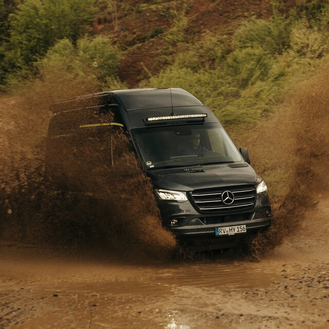 Camper Van fährt duch einen Offroad Park, er ist schmutzig, im Hintergrund sind Bäume und Dreckhügel zu sehen