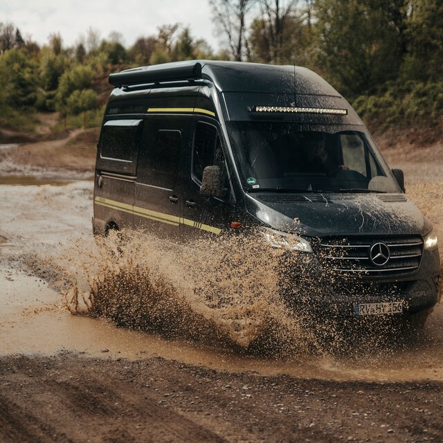 Camper Van fährt in der Ferne duch einen Offroad Park, im Hintergrund sind Bäume und Dreckhügel zu sehen und links neben dem Camper Van sieht man eine Pfütze