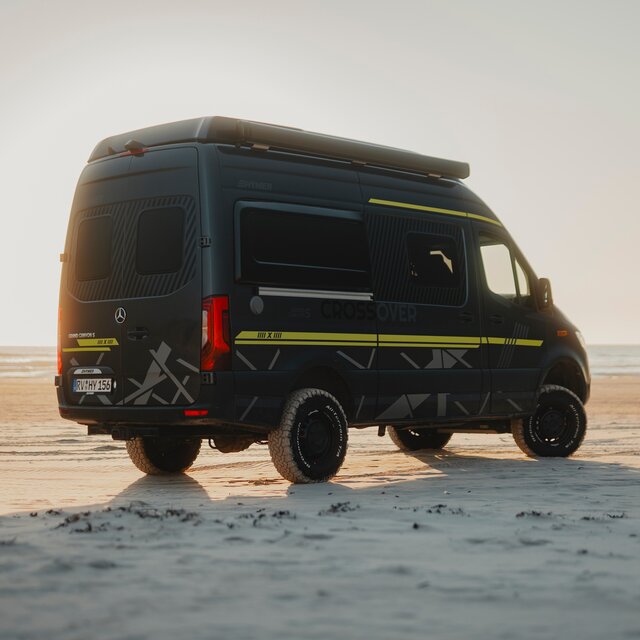 Ein Teil des schwarzen Kühlergrills eines schwarzen Camper Vans, eine graue 4x4 Abklebung sowie der rechte Frontscheinwerfer sind zu sehen, im Hintergrund sieht man Dünen