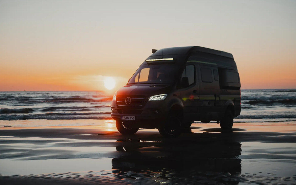 Schwarzer Camper Van mit Neongrüner Beklebung steht seitlich schräg an einem Strand, sodass man die Vorderseite sehen kann, im Hintergrund sieht man das Meer über dem die Sonne untergeht