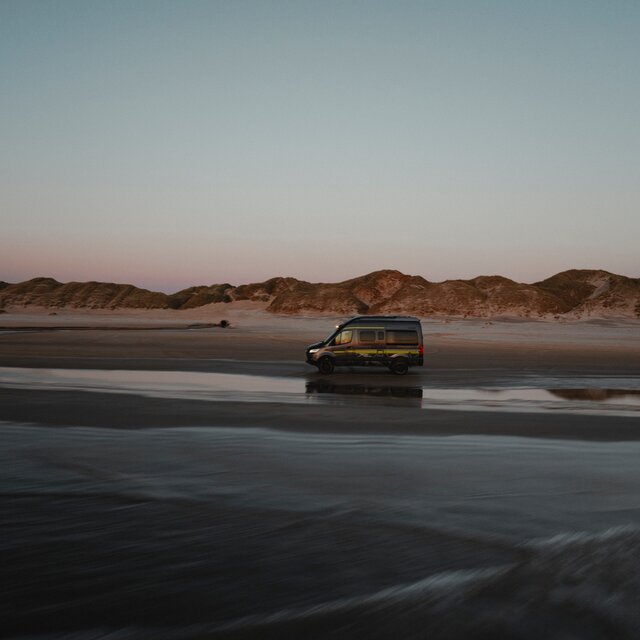 Schwarzer Camper Van mit Neongrüner Beklebung fährt seitlich über Sand, im Hintergrund sieht man Dünen