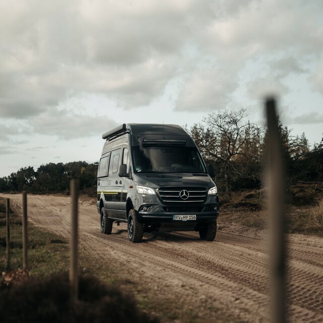 Schwarzer Camper Van fährt über eine matschige Straße, im Hintergrund sieht man Bäume und links neben der Straße sieht man einen Zaun mit Holzpflöcken