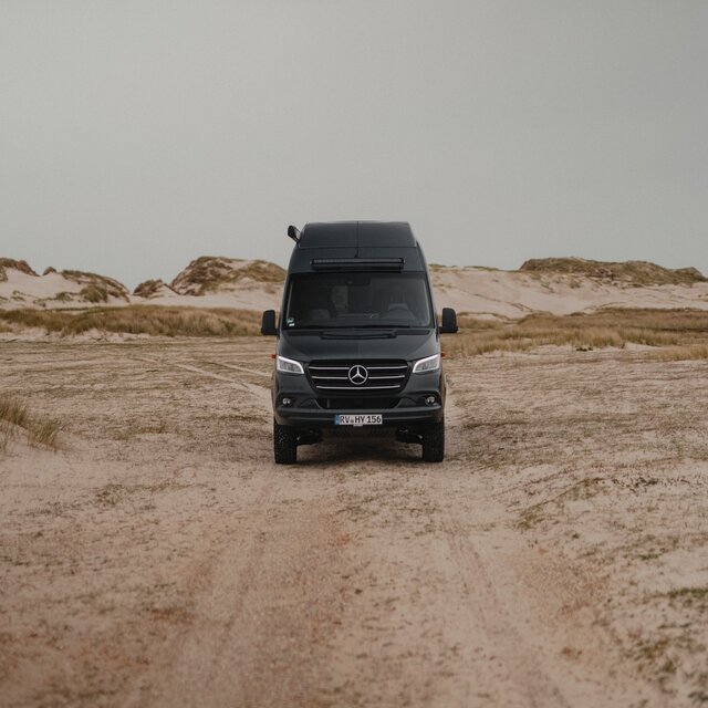 Schwarzer Camper Van von vorne, er steht auf Sand und im Hintergrund sieht man Dünen, über der Frontscheide sieht man eine LED-Lightbar