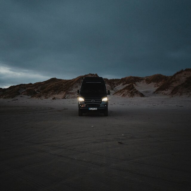 Schwarzer Camper Van von vorne, er steht auf Sand und im Hintergrund sieht man Dünen, es ist dunkel und die Scheinwerfer leuchten