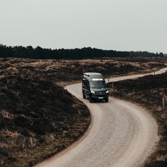 Schwarzer Camper Van fährt über eine steinige Straße, rechts und links von der Straße sieht man etwas höheres Gras, im Hintergrund ist ein Wald zu sehen, die Scheinwerfer und die LED-Lightbar oberhalb der Frontscheibe leuchten