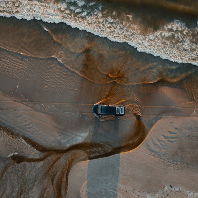 Schwarzer Camper Van fährt über den Strand, man sieht ihn von oben, er fährt parallel zum Meer