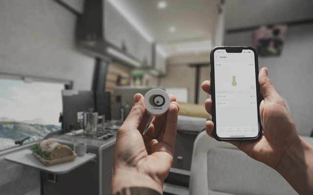 Deux mains au premier plan, dont l'une tient un capteur de température et l'autre un téléphone portable affichant une lecture de température. En arrière-plan, l'intérieur flou d'un camping-car est visible.