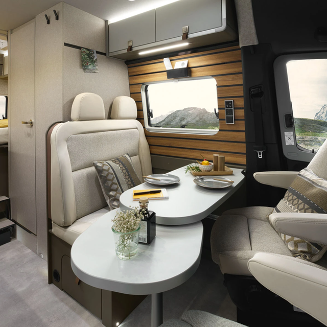 Intérieur moderne d'un camping-car avec des garnitures claires, montrant le coin salon avec une table de séjour pivotée. Le mur du coin salon est revêtu de panneaux en bois.