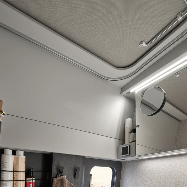 Gros plan sur une salle de bain de camping-car, montrant des compartiments de rangement avec des produits cosmétiques et à droite un grand miroir avec un miroir de maquillage rond fixé dessus.