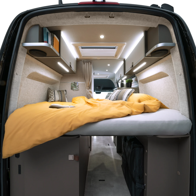 Vue à travers les portes arrière ouvertes dans un camping-car moderne. À l'arrière du véhicule se trouve le lit avec une literie jaune dessus.