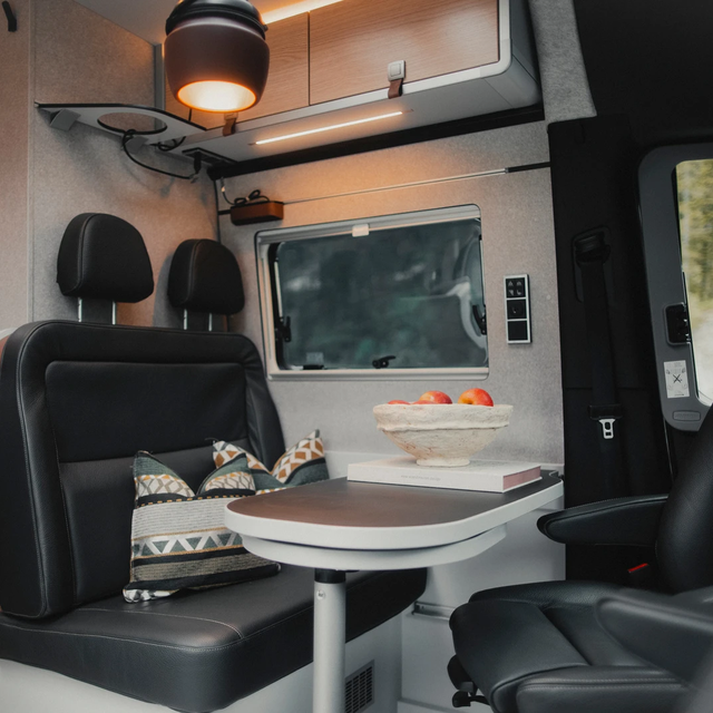 Espace de vie dans un fourgon aménagé moderne, table dressée avec coin salon en cuir sombre, les deux sièges conducteur sont tournés vers la table, au-dessus de la zone de sièges se trouvent des placards de rangement de toit et une lampe suspendue marron