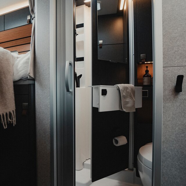 Bathroom in a motorhome with sink, cassette toilet, mirror and dark cabinets, next to it is the mattress of the sleeping area with blanket