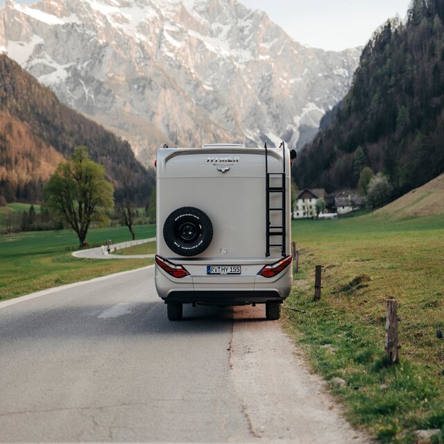 Motorhome from behind, standing on a road, mountains can be seen in the background, a black ladder and a black spare wheel can be seen on the rear of the vehicle