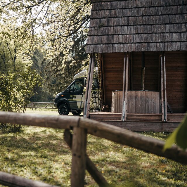 Side view of a dark driver's cab of a motorhome, protruding behind a wooden hut in a small forest
