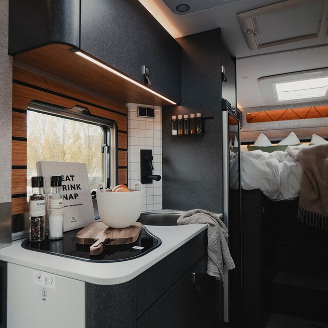 Interior of a motorhome showing the entrance door, kitchen unit with cooking utensils and worktop extension, refrigerator, in the background you can see the sleeping area of the motorhome with a blanket