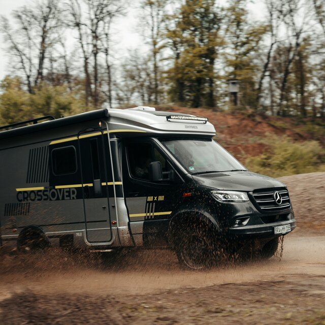 Semi-integrated motorhome driving through a small puddle in an off-road park, puddle splashes, trees and dirt mounds are visible in the background