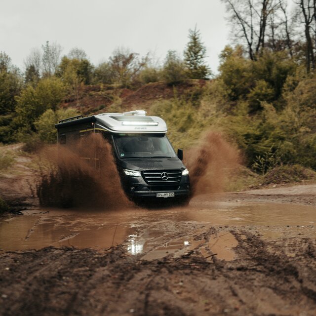 Semi-integrated motorhome driving through a large puddle in an off-road park, puddle splashes, trees and dirt mounds are visible in the background