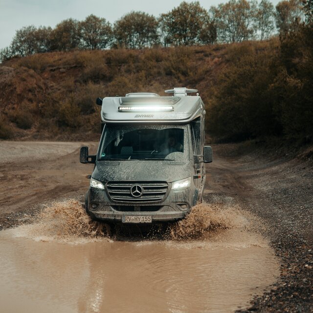 Semi-integrated 4x4 motorhome drives through a small puddle in an off-road park, trees and dirt mounds are visible in the background