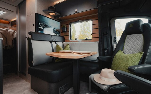 Modern living space in a motorhome, with driver seats, seating area, flat screen TV, side window, set table, dark leather decor, and overhead cabinet