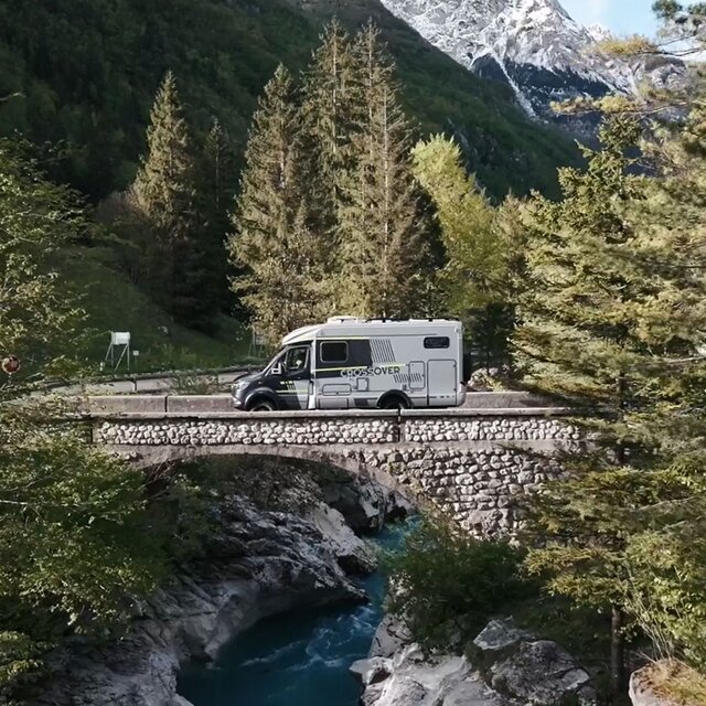 Semi-integrated motorhome drives over a bridge in the mountains, to the right and left of the bridge and in the background you can see trees, under the bridge flows a river