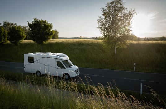 Side view of the moving HYMER Tramp S motorhome on a road next to a cycle path, grain fields and individual trees