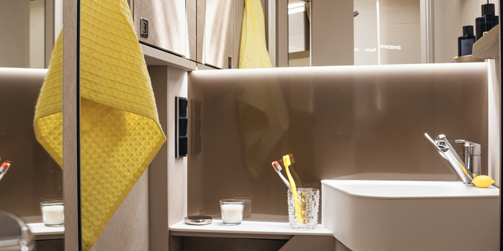 Spiegel, wastafel, opbergruimte, gele handdoek en toilet in de badkamer van de HYMER Tramp S camper