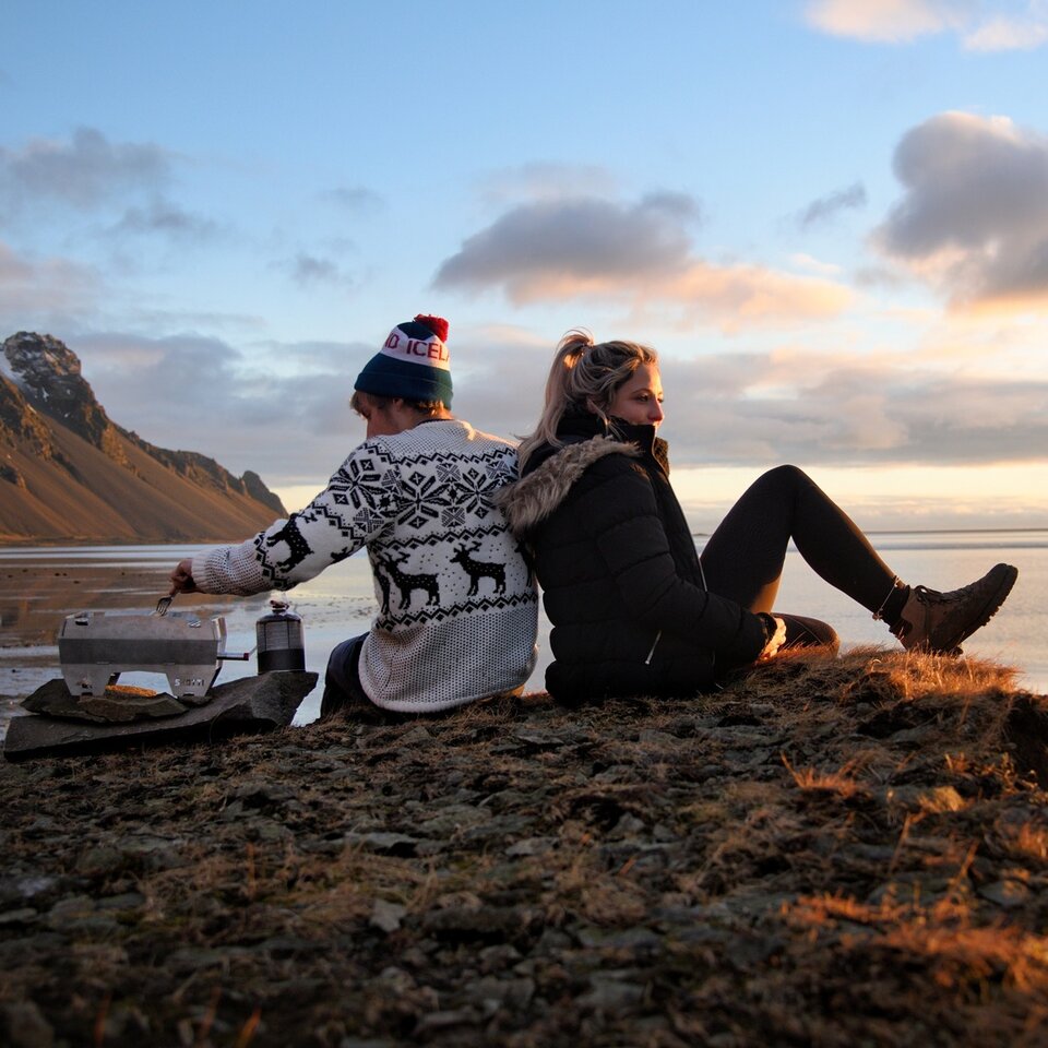 Två personer i vinterkläder sitter med grillen framför en vidsträckt utsikt med berg. Fotograferat under resor med husbil till Island.