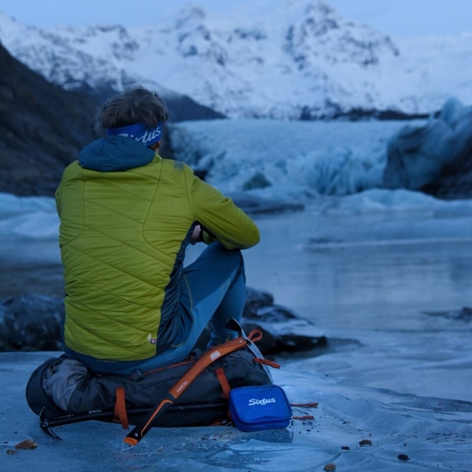Photo from a trip to Iceland by campervan. A man in a green jacket sits on a rucksack on a patch of ice with his back to the viewer.