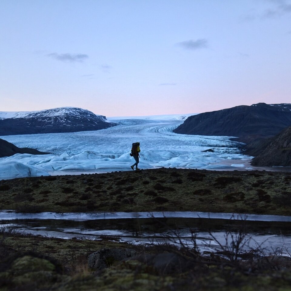 En person vandrar med ryggsäck genom ett landskap med gräs, kullar och snö. Bild från en reseberättelse till Island med husbil och färja.