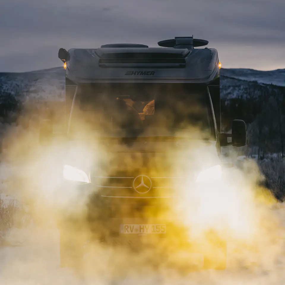 Blick von vorne auf den HYMER ML-T CrossOver mit eingeschalteten Scheinwerfern, Fahrzeug steht in einer Schneelandschaft, im Hintergrund sind Berge mit Schnee