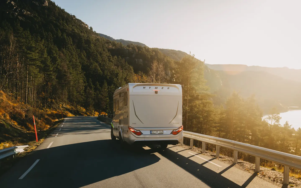 Blick von hinten auf auf den HYMER B-MC T BlackLine, Fahrzeug fährt auf einer Landstraße, Im Hintegrund links ist ein Wald, im Hintergrund rechts ist ein See