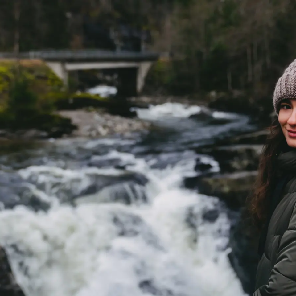 Blick auf eine Frau, im Hintergrund ist ein Fluss mit einem kleinen Wasserfall