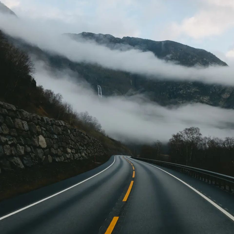 Blick auf eine Straße, die umgeben ist von Gebirge, im Hintergrund ist Nebel zu sehen