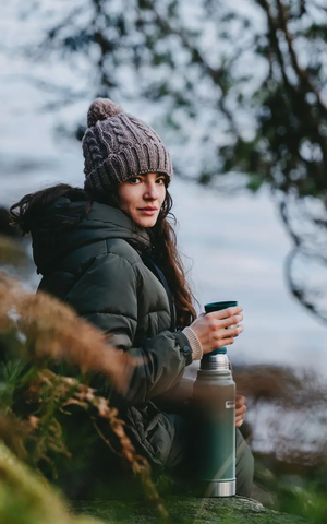 Eine warm bekleidete Frau mit Isolierflasche sowie einem Becher in der Hand sitzt auf einem Felsen in freier Natur