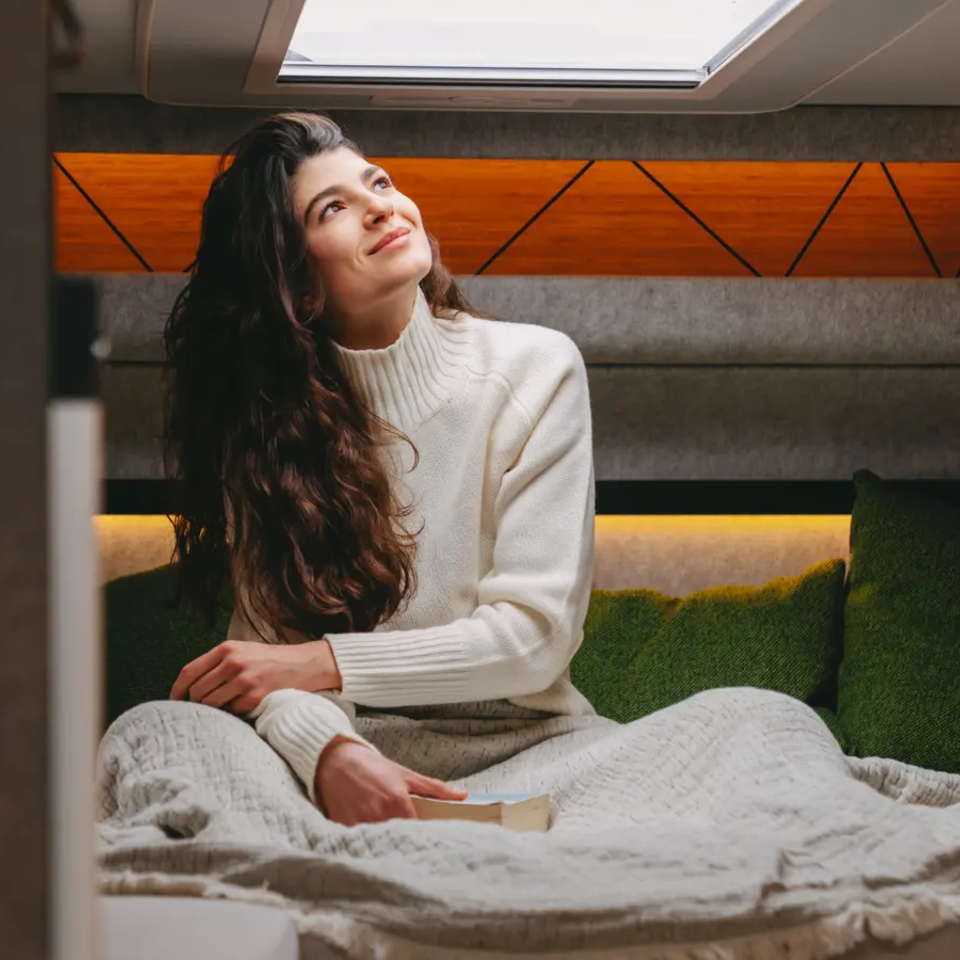 Eine Frau sitzt im Scheidersitz im Heckbett eines modernen Wohnmobils und schaut aus dem Dachfenster