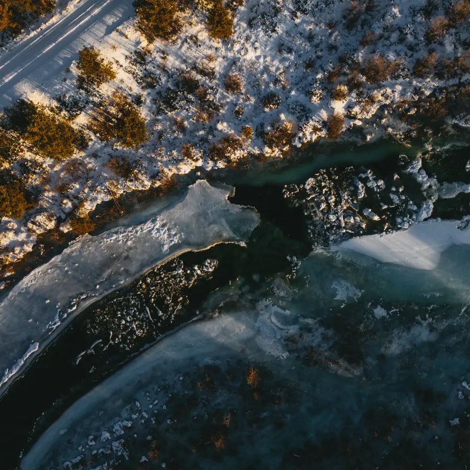 Luftblick auf einen fast vereisten Fluss, daneben ist eine verschneite Straße zu sehen