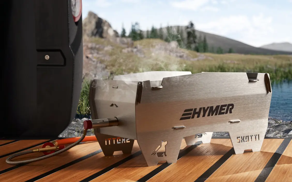 Skottigrill steht auf Holzdielen, im Hintergrund ist ein Gewässer und hügelige Landschaft zu sehen