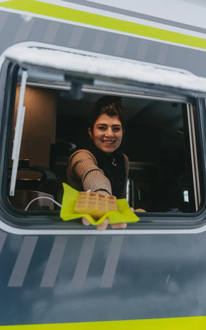 Blick in das Küchenfenster des HYMER ML-T 570 CrossOver, aus der eine Frau eine Waffel herausreicht