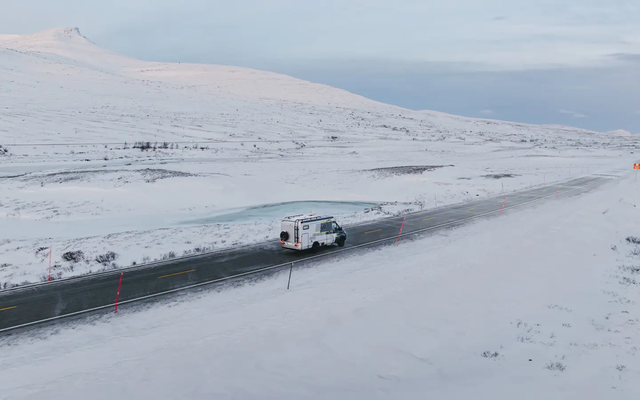 Seitlicher Luftblick auf die Beifahrerseite des HYMER B-MC T BlackLine, der eine Straße langfährt, im Hintergrund sind Schneeberge