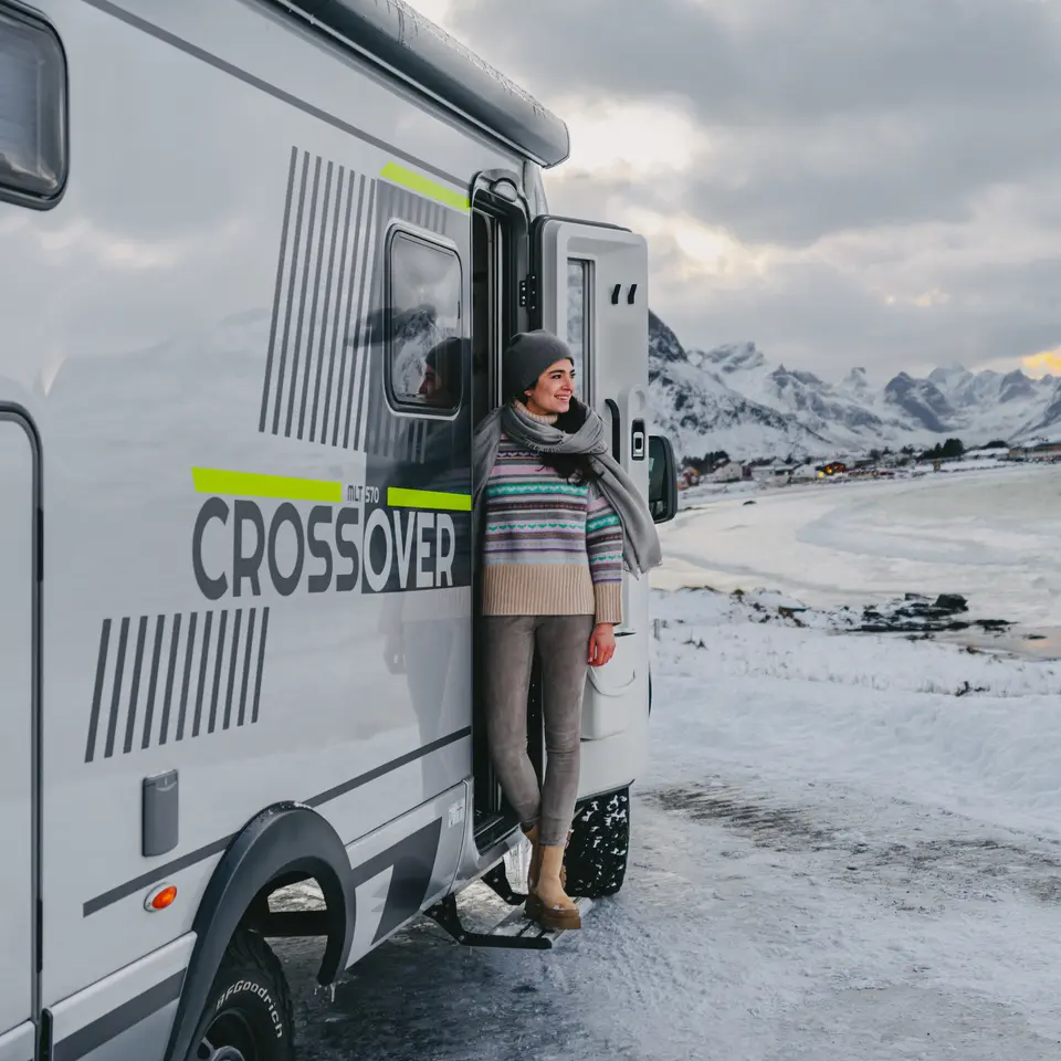 Frau steht an der Eingangstür vom HYMER ML-T 570 CrossOver, im Hintergrund ist ein kleines Dorf an einer Meeresbucht